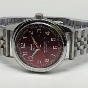 May include: A silver-toned wristwatch with a burgundy dial and matching hands. The watch face displays numerals and the text "PILOT" and "PARA SHOCK". The watch has a silver link band.