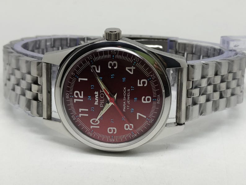 May include: A silver-toned wristwatch with a burgundy dial and matching hands. The watch face displays numerals and the text "PILOT" and "PARA SHOCK". The watch has a silver link band.