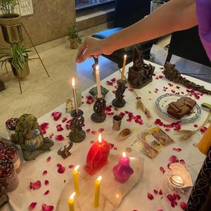 Op de afbeelding: Een witte tafel bedekt met verspreide rode rozenblaadjes, meerdere brandende kaarsen in verschillende kleuren, een rode hartvormige kaars, een paarse figuurkaars, tarotkaarten, een bordje met een stukje cake en decoratieve beeldjes.
