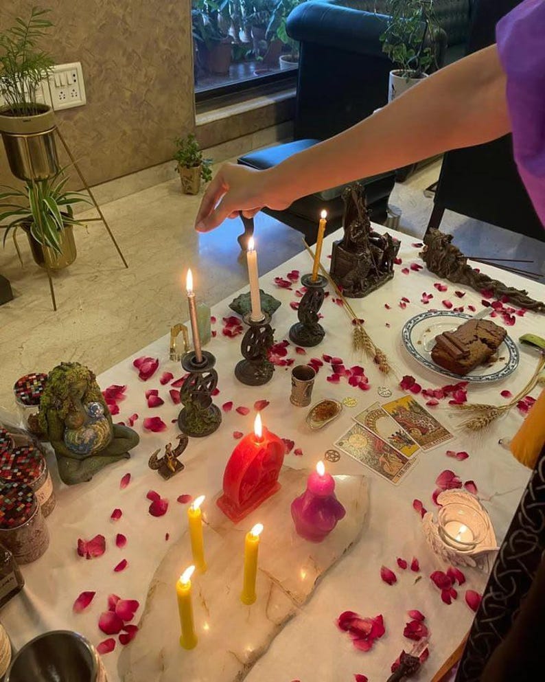 Op de afbeelding: Een witte tafel bedekt met verspreide rode rozenblaadjes, meerdere brandende kaarsen in verschillende kleuren, een rode hartvormige kaars, een paarse figuurkaars, tarotkaarten, een bordje met een stukje cake en decoratieve beeldjes.