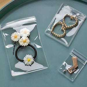 Könnte beinhalten: Transparente Zip-Top-Beutel mit Schmuck und Haaraccessoires. Eine Tasche enthält ein schwarzes Haargummi mit Gänseblümchen-Akzenten. Eine andere enthält goldene herzförmige Ohrringe. Eine dritte Tasche enthält goldene Ohrringe.