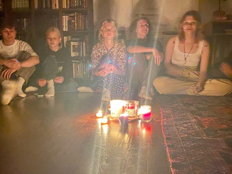 Op de afbeelding: Vijf personen zitten in een cirkel op een donkere vloer rond een verzameling brandende kaarsen in glazen potten. De groep bevindt zich voor een hoge houten boekenkast vol boeken. De warme gloed van de kaarsen verlicht de hele sc&egrave;ne.
