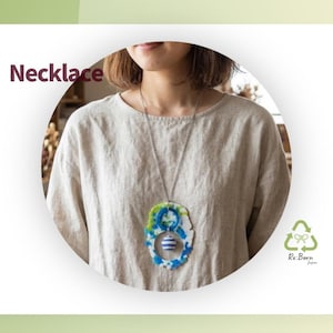 Puede incluir: Un collar con una cadena larga de tono plateado y un colgante grande e irregular de material jaspeado en azul, verde y blanco. El colgante incluye un recorte circular que sostiene una pequeña cuenta esférica con rayas azules y blancas.