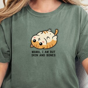 Może przedstawiać: Zielona koszulka w kolorze mchu z ilustracją kota z kreskówki. Kot jest pomarańczowo-biały, leży na plecach z napisem "MAMA, I AM BUT SKIN AND BONES" poniżej. Koszulka ma luźny krój.