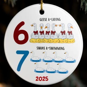 May include: A white, round Christmas ornament with a gold string. The ornament features the numbers 6 and 7, geese and swans illustrations, and the year 2025. Text includes "Geese A-Laying" and "Swans A-Swimming."