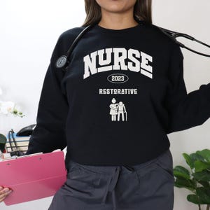 May include: Black sweatshirt with the word "NURSE" in white block letters, the year "2023", and the word "RESTORATIVE" below. A white graphic of a nurse assisting a patient is also featured. A stethoscope is around the neck.
