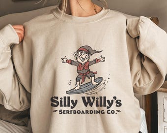 Könnte beinhalten: Beiges Sweatshirt mit einem Cartoon-Elf, der auf einem grauen Surfbrett surft. Der Elf trägt eine rote Weste und einen Hut. Der Text "Silly Willy's SERFBOARDING CO." ist darunter in Dunkelbraun aufgedruckt.