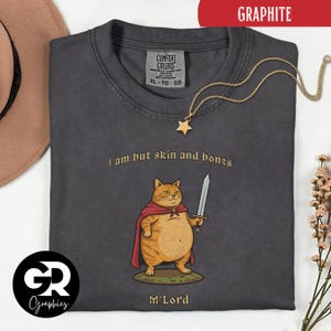 Może przedstawiać: Grafitowa szara koszulka z ilustracją kota z kreskówki. Kot, w czerwonej pelerynie i trzymający miecz, jest oznaczony jako "M'Lord". Tekst "I am but skin and bones" znajduje się nad kotem. Złoty naszyjnik z wisiorkiem w kształcie gwiazdy jest zarzucony na koszulkę.