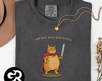 May include: Graphite grey t-shirt featuring a cartoon cat illustration. The cat, wearing a red cape and holding a sword, is labelled "M'Lord." The text "I am but skin and bones" is above the cat. A gold star pendant necklace is draped on the shirt.