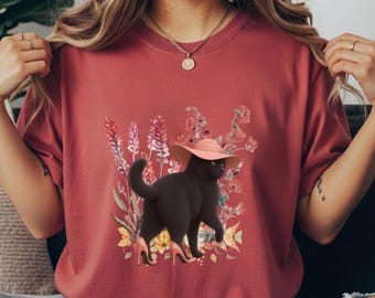 Può includere: T-shirt bordeaux con un'illustrazione stravagante di un gatto nero con cappello rosa e tacchi alti, su uno sfondo di fiori colorati. Il gatto è il fulcro centrale, aggiungendo un tocco giocoso al design.