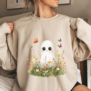 Puede incluir: Sudadera beige con un diseño de fantasma blanco rodeado de flores y mariposas. El fantasma tiene ojos grandes, creando un aspecto lindo y caprichoso. La sudadera tiene cuello redondo y mangas largas.
