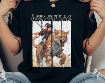 May include: Black t-shirt featuring an illustration of a person riding a tiger. The tiger is orange with black stripes and has an open mouth. The person is wearing armor and a red cape. Text on the shirt reads "Always land on my feet, always protect my liege."