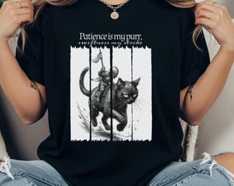 May include: Black t-shirt featuring a graphic of a knight riding a black cat, with the text "Patience is my purr, swiftness my strike." The design is in black and white, with vertical lines behind the cat and knight.