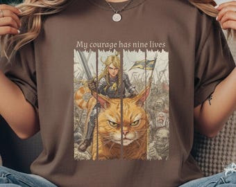 May include: Brown t-shirt featuring a graphic of a warrior riding a large orange cat, holding a sword and a flag. The text "My courage has nine lives" is above the image. The design has a fantasy theme.