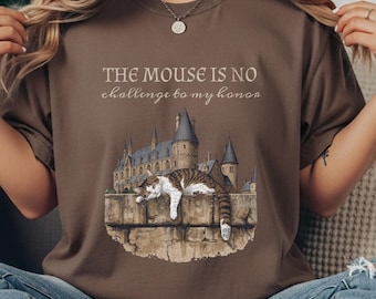 Puede incluir: Camiseta marrón con un gráfico de un gato descansando sobre un muro de piedra frente a un castillo. El texto "THE MOUSE IS NO challenge to my honor" está impreso encima del gráfico. La camiseta está hecha de un material suave.