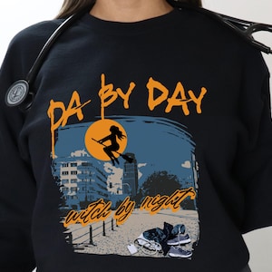 Può includere: Felpa nera con la scritta arancione "PA by DAY witch by night". La grafica presenta una strega su una scopa davanti a una luna piena, con uno skyline e delle scarpe da ginnastica a terra.