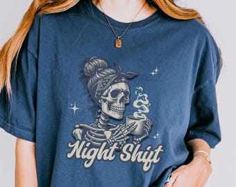 以下が含まれることがあります： ネイビーブルーのTシャツ。バンダナを巻いた骸骨がコーヒーカップを持ち、「Night Shift」の文字がヴィンテージフォントで描かれています。デザインには湯気と星のアクセントが含まれています。