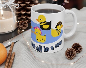 Puede incluir: Taza de cerámica blanca con tres patos de dibujos animados con accesorios punk y moteros sobre un diseño de olas azules. La taza tiene el texto "not all in a row". La taza está llena de un líquido oscuro.