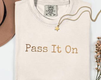 Puede incluir: Camiseta color crema con la frase bordada "Pass It On" en color dorado. También se ven un collar con colgante de estrella dorada y un sombrero marrón. La camiseta tiene una etiqueta que dice "Comfort Colors".