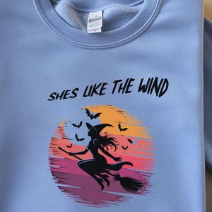 Può includere: Felpa azzurra con la scritta "Shes Like The Wind" sopra una grafica di una strega che vola su una scopa davanti a un tramonto con pipistrelli. La strega indossa un cappello.