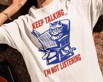 May include: White t-shirt with a blue and red graphic of a cat wearing glasses and reading a newspaper in a chair. The text "KEEP TALKING.. I'M NOT LISTENING" is printed above and below the cat illustration.
