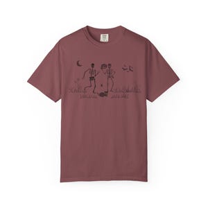 Könnte beinhalten: Burgunderfarbenes T-Shirt mit schwarzem Grafikdesign. Das Design zeigt zwei Skelette, eines mit einem Kürbis, sowie einen Mond, Fledermäuse, eine Spinne und eine Katze. Ein bequemes, lässiges Kleidungsstück.