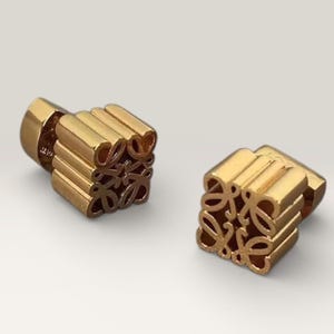 May include: Two gold-colored cube-shaped earrings. Each earring features a repeating pattern of a stylized floral design. The earrings are made of a shiny, metallic material and are set against a neutral background.