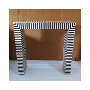 May include: A modern console table with a black and white striped pattern. The table has a rectangular top and two legs, each with a matching striped design. The table is made of wood and is approximately 30 inches wide.