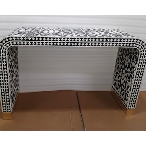 May include: A black and white patterned console table with a curved design. The table features a geometric pattern and gold-colored accents on the base. The table's dimensions are approximately 48 inches wide, 16 inches deep, and 30 inches high.