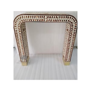 May include: A decorative console table with a U-shaped design. The table features a white background with a brown leaf pattern. The legs and base are a gold color. The table is approximately 30 inches wide.