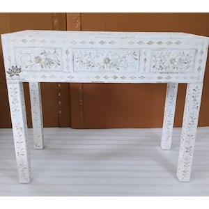 May include: White wooden desk with three drawers and four legs. The desk is decorated with floral and leaf patterns. The legs and drawers are adorned with inlaid mother-of-pearl accents. The desk is a decorative piece of furniture.