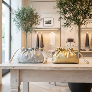 May include: Two drawstring pouches on a wooden table. One is light blue with a floral pattern, the other is gold. Both have cream-colored drawstrings. The background shows a clothing store with racks of clothing.