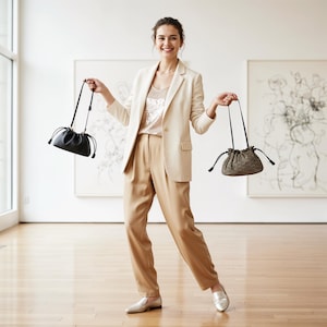 May include: A woman wearing a cream-colored blazer, tan pants, and silver shoes, holding two drawstring handbags. One bag is black, and the other is a patterned brown. The background features abstract art and a wooden floor.