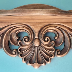 May include: Close-up of a wooden decorative shelf with ornate, carved scrollwork and a central shell-like design. The shelf is stained in a warm brown tone, with a light brown shelf area. The background is a solid teal color.