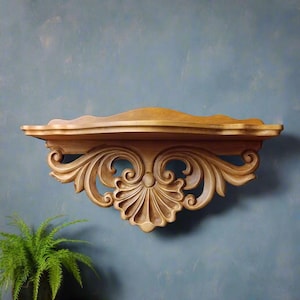 May include: A wooden wall shelf with ornate, carved details. The shelf is a light brown color and features a scalloped edge and intricate scrollwork. The design includes a central fan-shaped element and symmetrical flourishes, perfect for displaying decorative items.