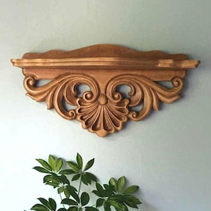 May include: A decorative wooden wall shelf with ornate, carved details. The shelf is a warm, golden brown color and features a scalloped top edge and intricate scrollwork. The shelf is approximately 24 inches wide.