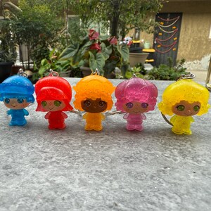 May include: Five colorful resin keychains in blue, red, orange, pink, and yellow. Each keychain features a small, stylized figure with glitter accents, a round head, and a silver keyring.