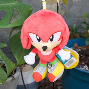 May include: A plush toy of Knuckles the Echidna, a red echidna character from the Sonic the Hedgehog franchise. The toy has white gloves, yellow and green shoes, and a tag attached. The toy is hanging from a beaded string.
