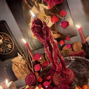 May include: A deep red wax candle sculpted into the form of two figures embracing, set upon a circular base. The candle is surrounded by scattered red rose petals, sliced apples, and lit red taper candles, with a Baphomet illustration in the background.