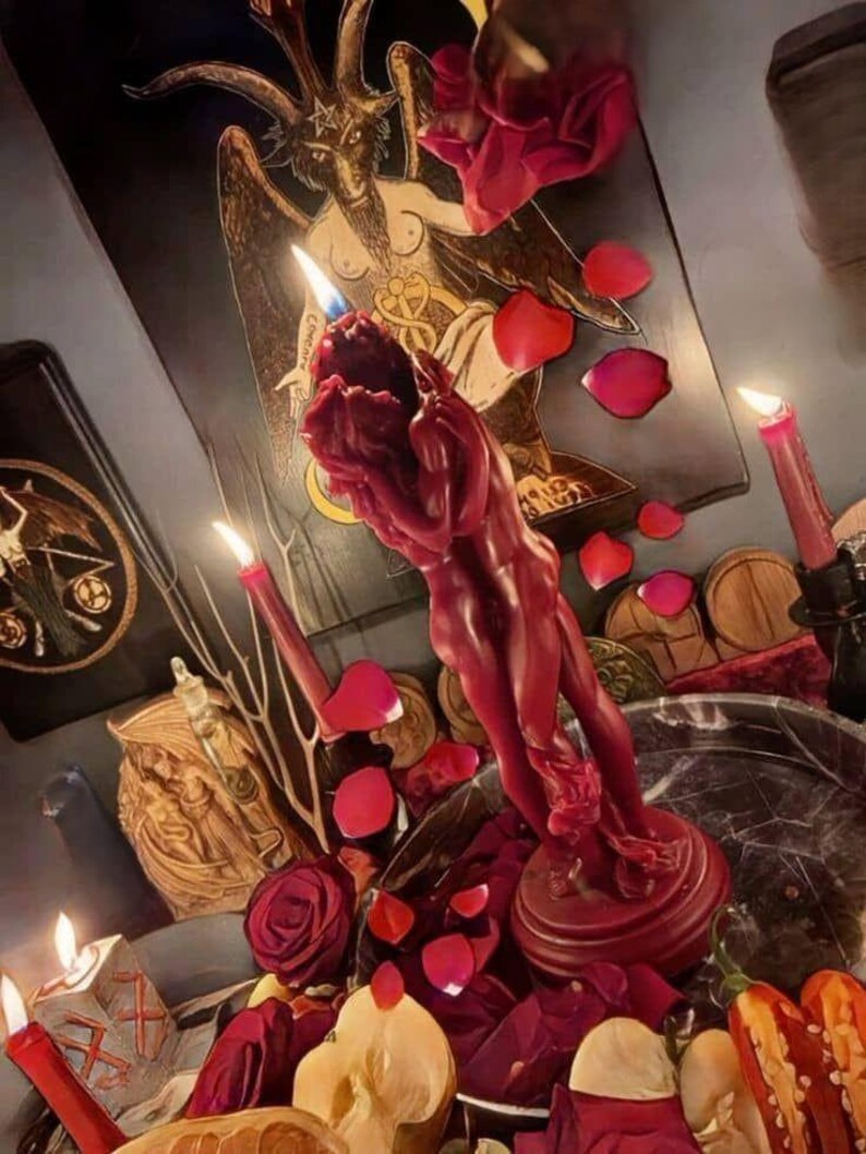 May include: A deep red wax candle sculpted into the form of two figures embracing, set upon a circular base. The candle is surrounded by scattered red rose petals, sliced apples, and lit red taper candles, with a Baphomet illustration in the background.