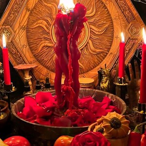 May include: A red candle shaped like two figures embracing, topped with a flame, set in a bowl filled with red rose petals. The scene includes red taper candles, fresh strawberries, red apples, and a large red rose in the foreground.