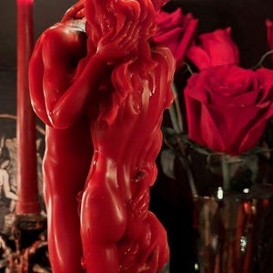 May include: A bright red wax candle sculpted into the shape of two figures embracing. The candle features a flame at the top, with detailed textures representing the forms of the figures. The wax is smooth and vibrant, capturing the figures in a close pose.
