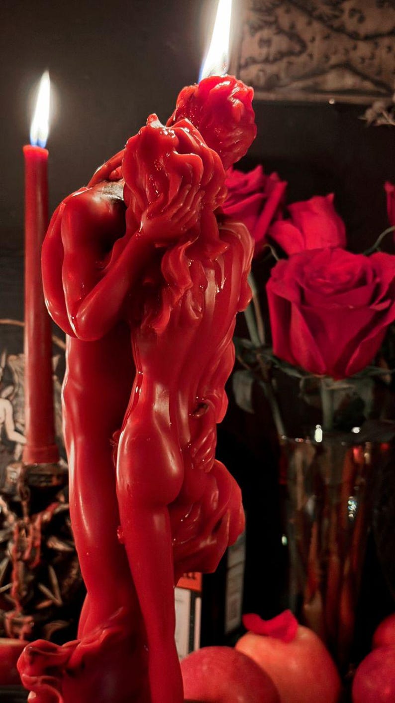 May include: A bright red wax candle sculpted into the shape of two figures embracing. The candle features a flame at the top, with detailed textures representing the forms of the figures. The wax is smooth and vibrant, capturing the figures in a close pose.