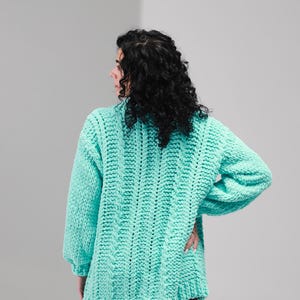 May include: A chunky knit sweater in a vibrant mint green color featuring a prominent vertical ribbed texture, long sleeves with dropped shoulders, and a relaxed, oversized fit that extends to the upper thigh area.