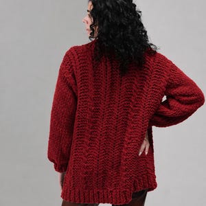 May include: A deep red, chunky knit jumper featuring a prominent vertical cable pattern throughout the body and sleeves. The garment has long, loose-fitting sleeves with ribbed cuffs and a thick, textured knit construction with a high neckline.