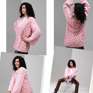 May include: A chunky knit cardigan in a soft pink hue featuring a prominent cable knit pattern throughout the body and sleeves. The garment has long sleeves, an open front design, and a relaxed fit. The text CARDI MINI love is visible.