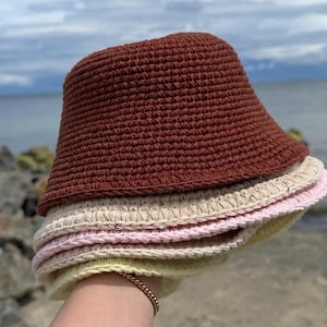 Op de afbeelding: Een stapel van vier gehaakte bucket hats in effen kleuren. De bovenste hoed is donkerbruin, gevolgd door een crèmekleurige hoed, een lichtroze hoed en een lichtgele hoed onderaan. Elke hoed heeft een ronde kroon en een korte rand.
