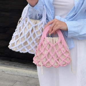 May include: Two handmade crochet bags. One is white, the other is pink. Both have a diamond-patterned net design over a beige fabric lining. The handles are made of the same crochet material. The bags are small, perfect for carrying essentials.