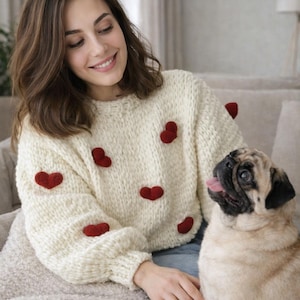 May include: A cream-coloured, chunky knit jumper featuring a crew neckline, long sleeves with ribbed cuffs, and several small, raised red heart-shaped appliqués scattered across the front and sleeves of the garment.