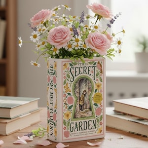 Könnte beinhalten: Ein Blumenarrangement in einer Vase in Form eines Buches mit dem Titel "The Secret Garden". Die Vase ist mit rosa Rosen, weißen Gänseblümchen und Lavendel gefüllt. Die Buchvase steht auf einem Holztisch mit anderen Büchern.